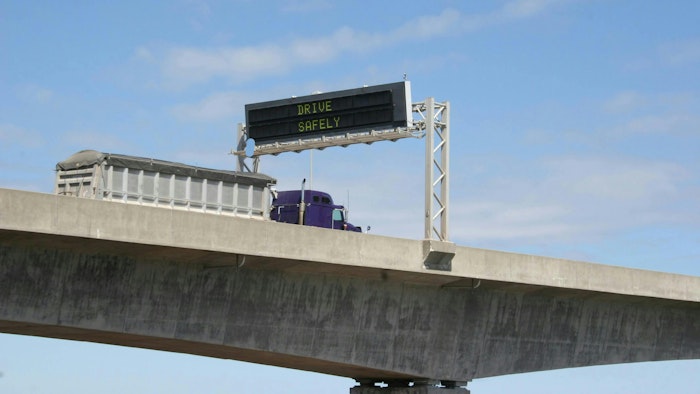 A tractor-trailer drives under a 'Drive Safely' display on a highway bridge. Operation Safe Driver Week returns to the highways across North America in July 2024.