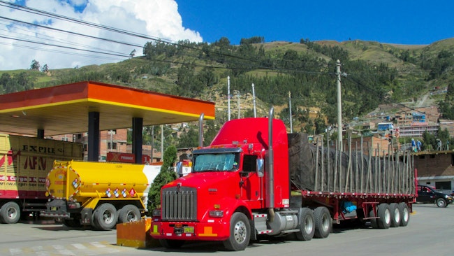Red semi-truck with flatbed trailer parked at a fuel station in a mountainous town, highlighting freight transport and trucking industry operations.