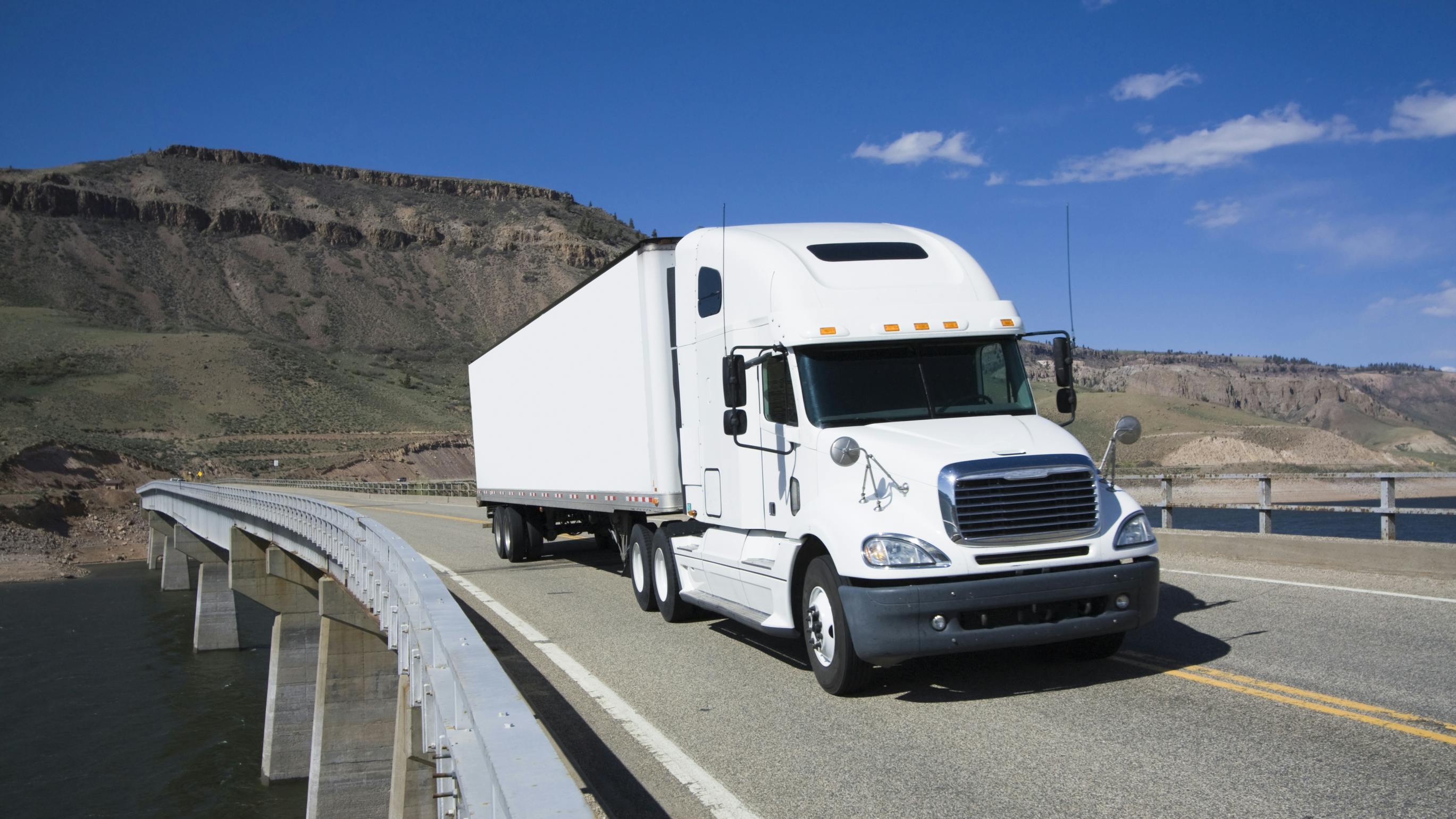 White semi truck with trailer driving across a highway bridge in a scenic mountain landscape under a clear blue sky.