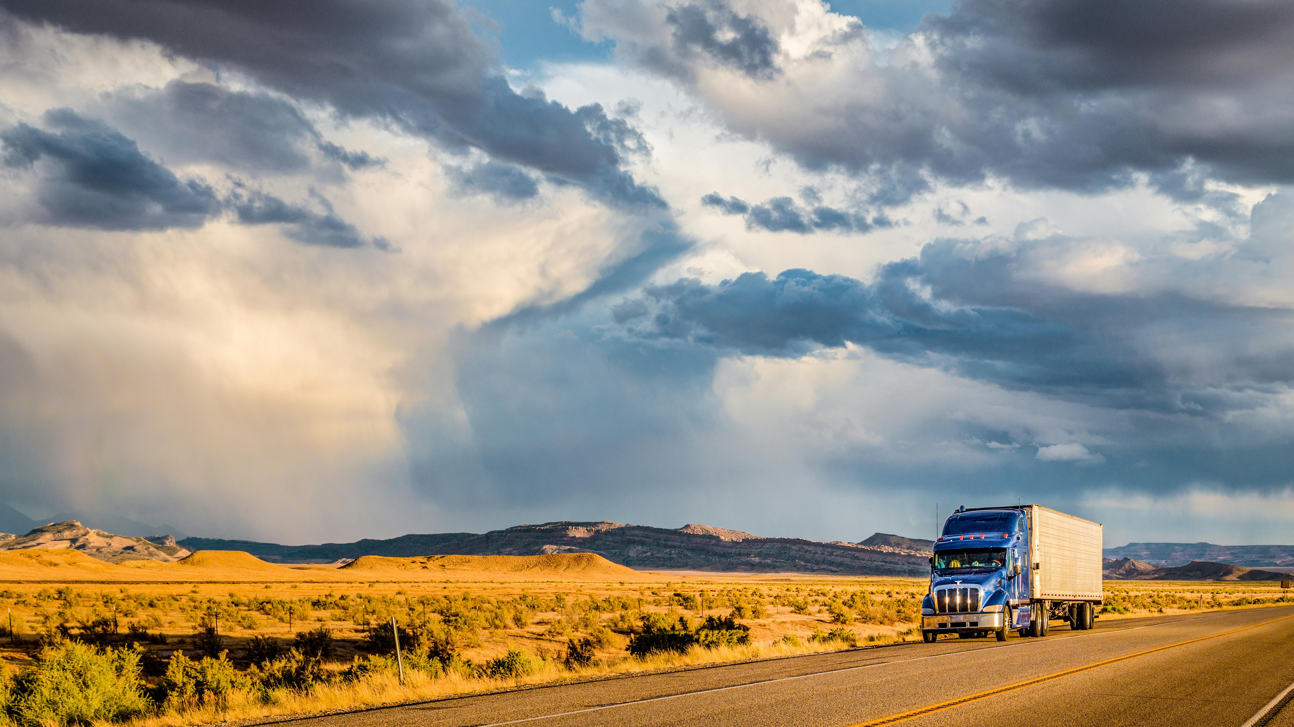 Blue semi-truck with trailer driving on a highway through the desert under dramatic storm clouds, symbolizing long-haul trucking and freight transport.