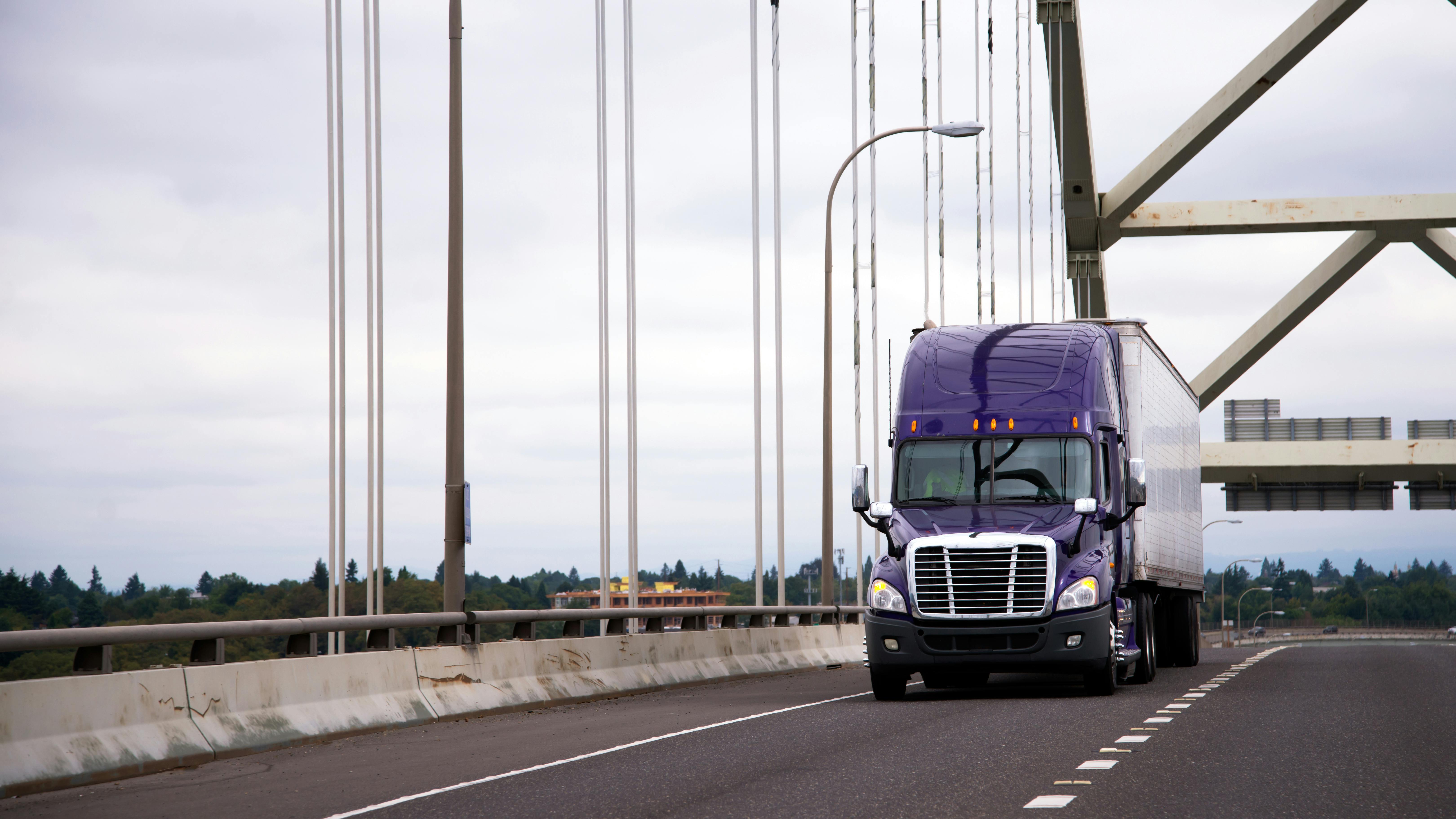 Purple semi-truck with a trailer crossing a highway bridge on a cloudy day, representing freight transportation and the trucking industry.