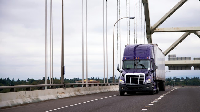 Purple semi-truck with a trailer crossing a highway bridge on a cloudy day, representing freight transportation and the trucking industry.