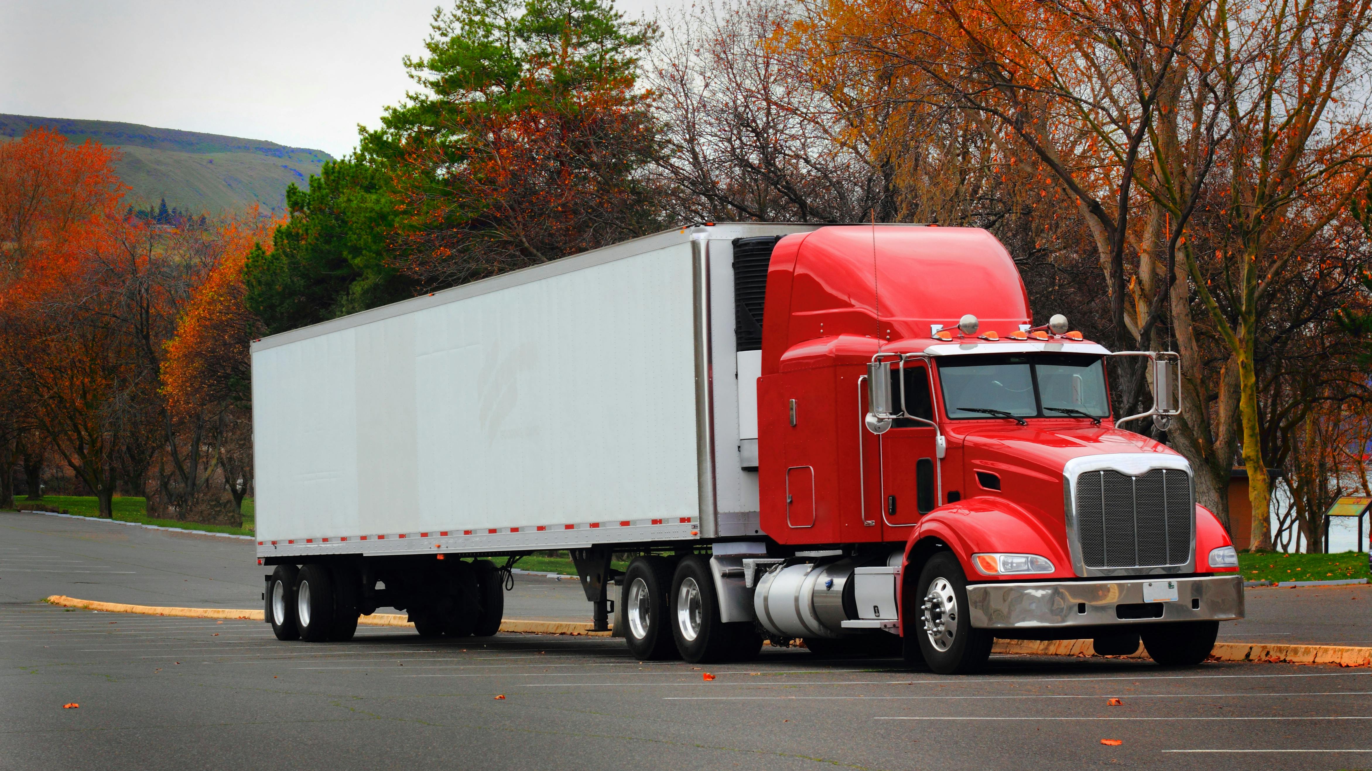 Red cab semi-truck with white trailer parked in a lot, side view under overcast skies with autumn-colored trees in the background.