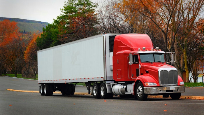 Red cab semi-truck with white trailer parked in a lot, side view under overcast skies with autumn-colored trees in the background.