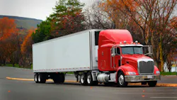 Red cab semi-truck with white trailer parked in a lot, side view under overcast skies with autumn-colored trees in the background. Red cab semi-truck with white trailer parked in a lot, side view under overcast skies with autumn-colored trees in the background.