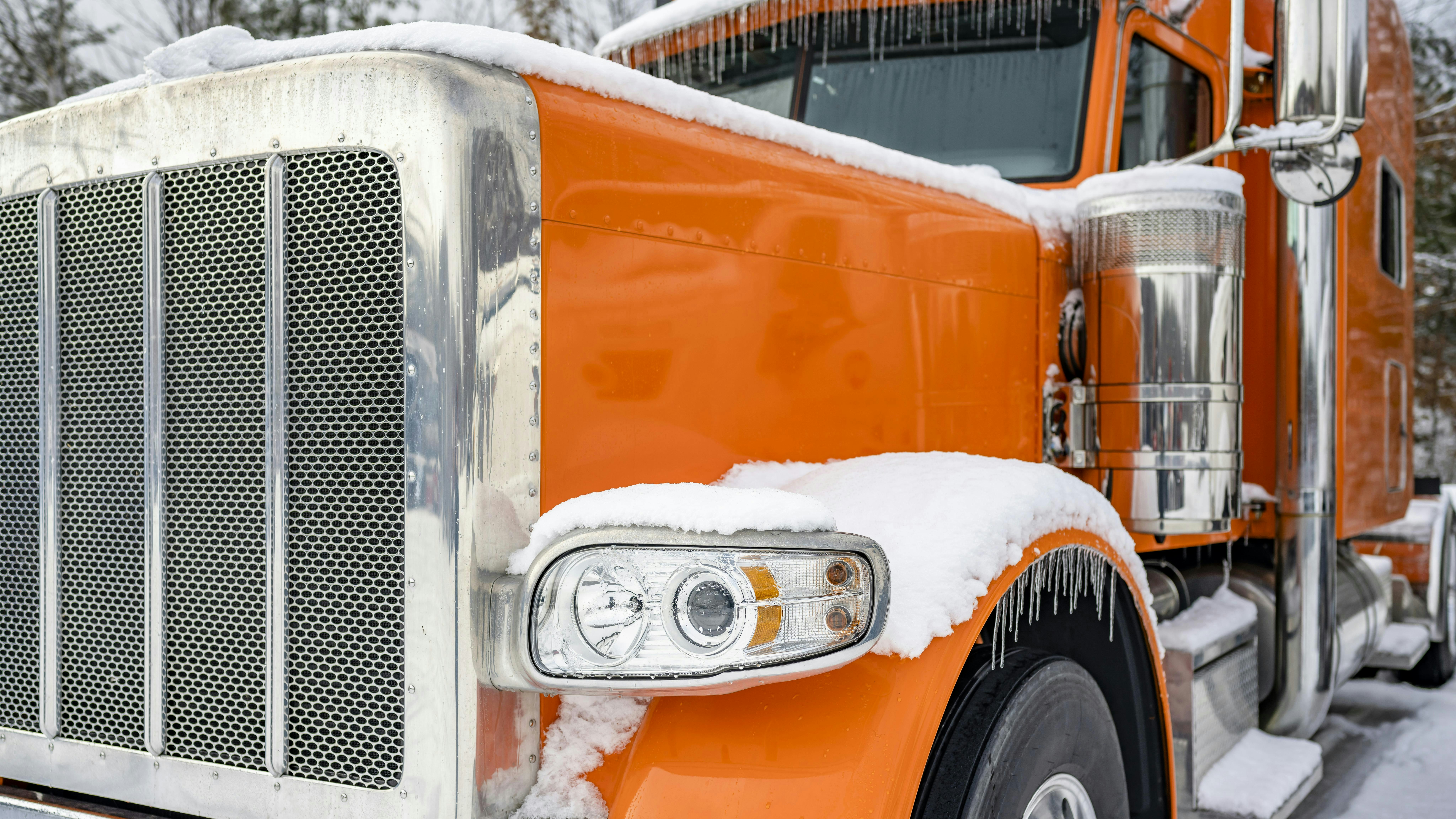 A classic orange big rig semi-truck with an extended cab parked in a snowy and icy truck stop parking lot during winter.