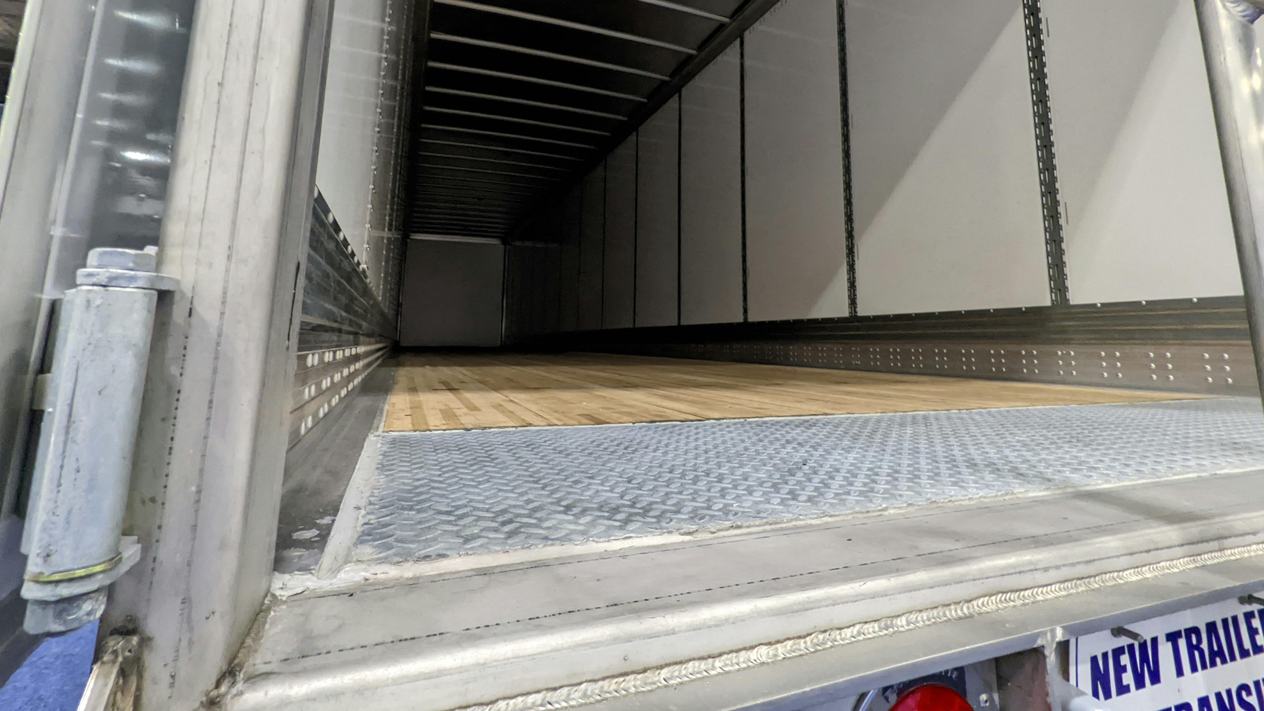 A view looking into the empty cargo space of a new commercial dry van trailer from the open rear doors, featuring a wooden floor, metal interior walls, and a diamond plate threshold.