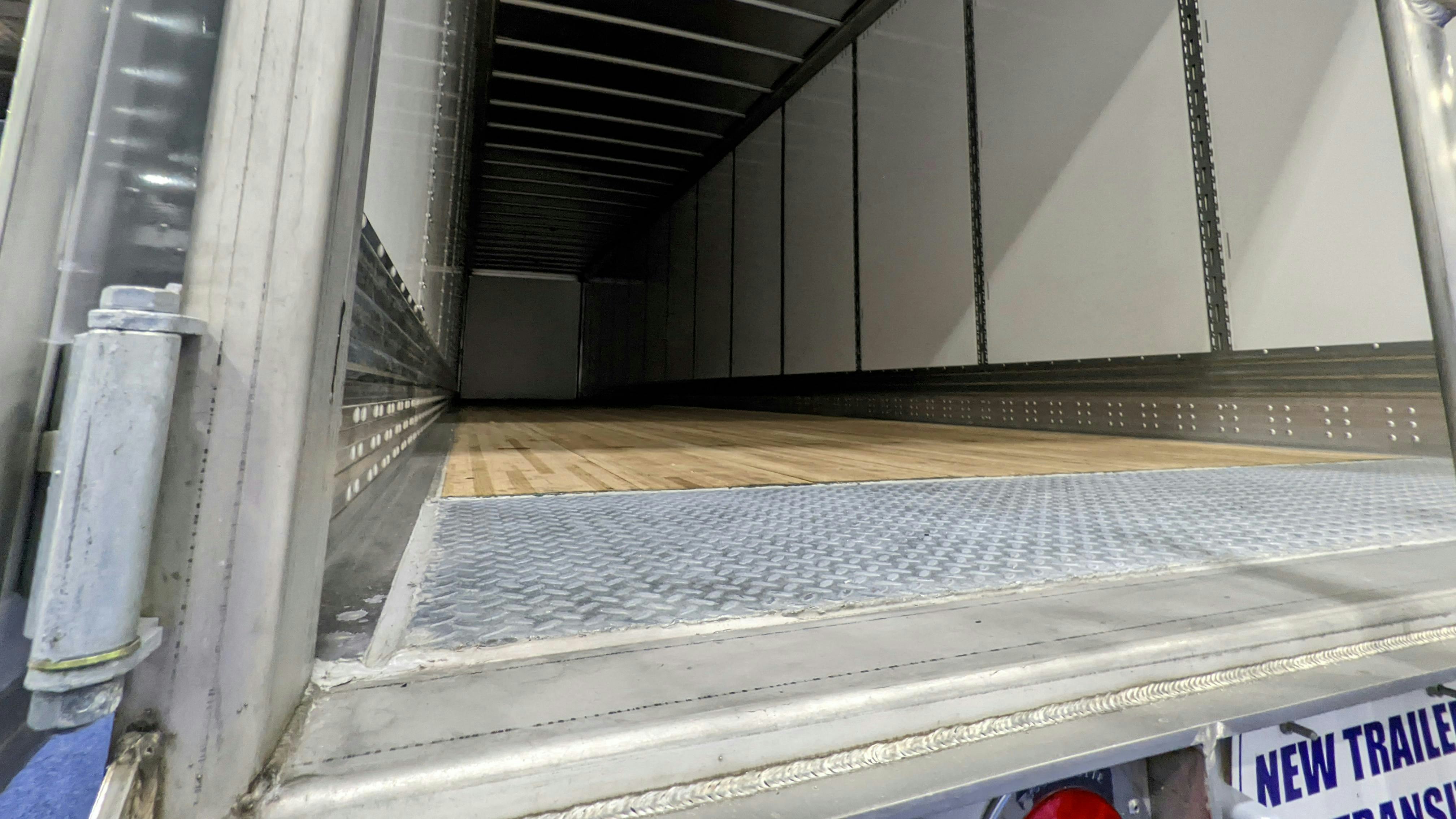 A view looking into the empty cargo space of a new commercial dry van trailer from the open rear doors, featuring a wooden floor, metal interior walls, and a diamond plate threshold.