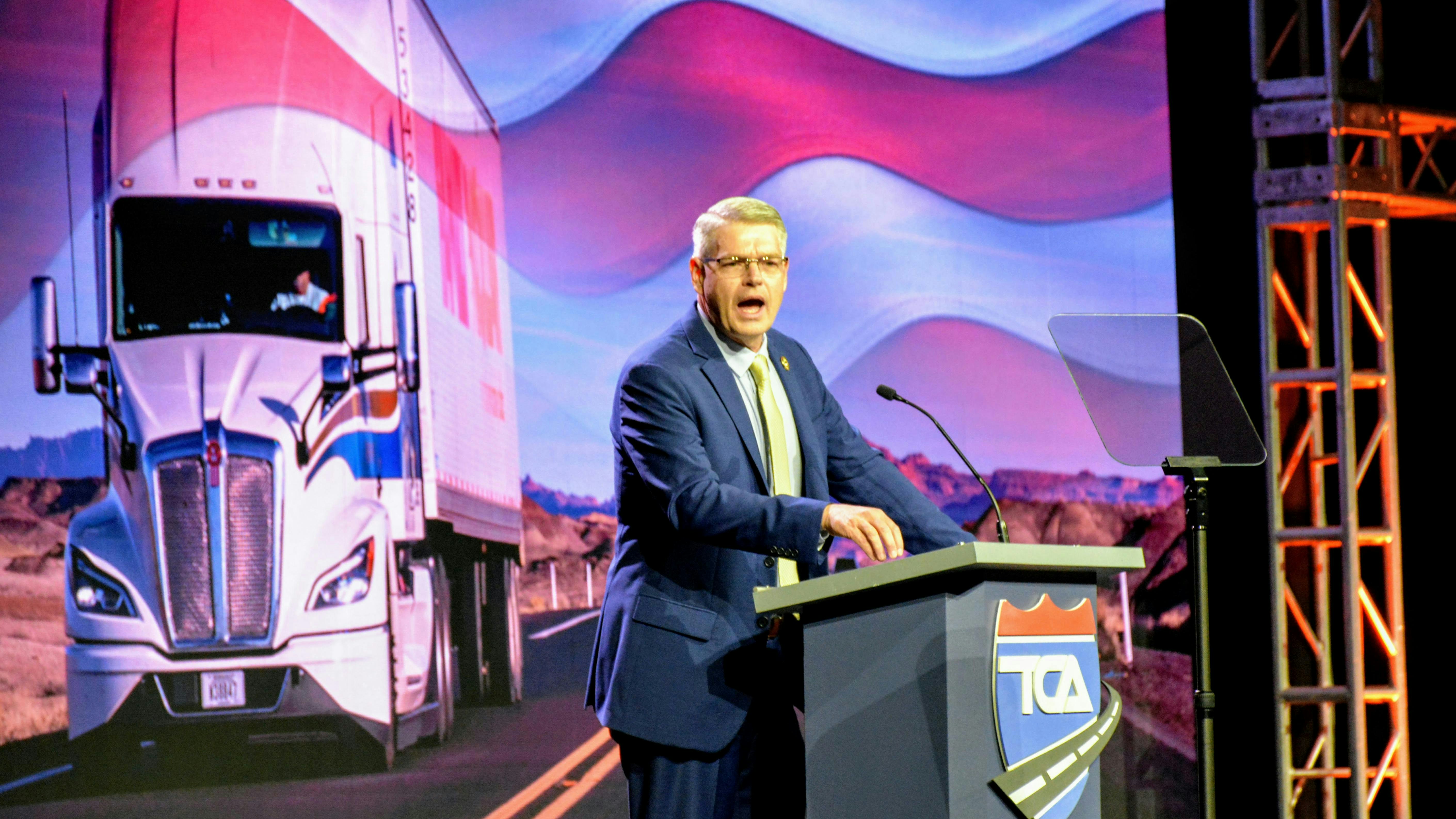 FMCSA Administrator Derek Barrs speaking passionately at a podium with the TCA logo during the Truckload 2026 convention, with a large screen displaying a semi-truck and American flag behind him.