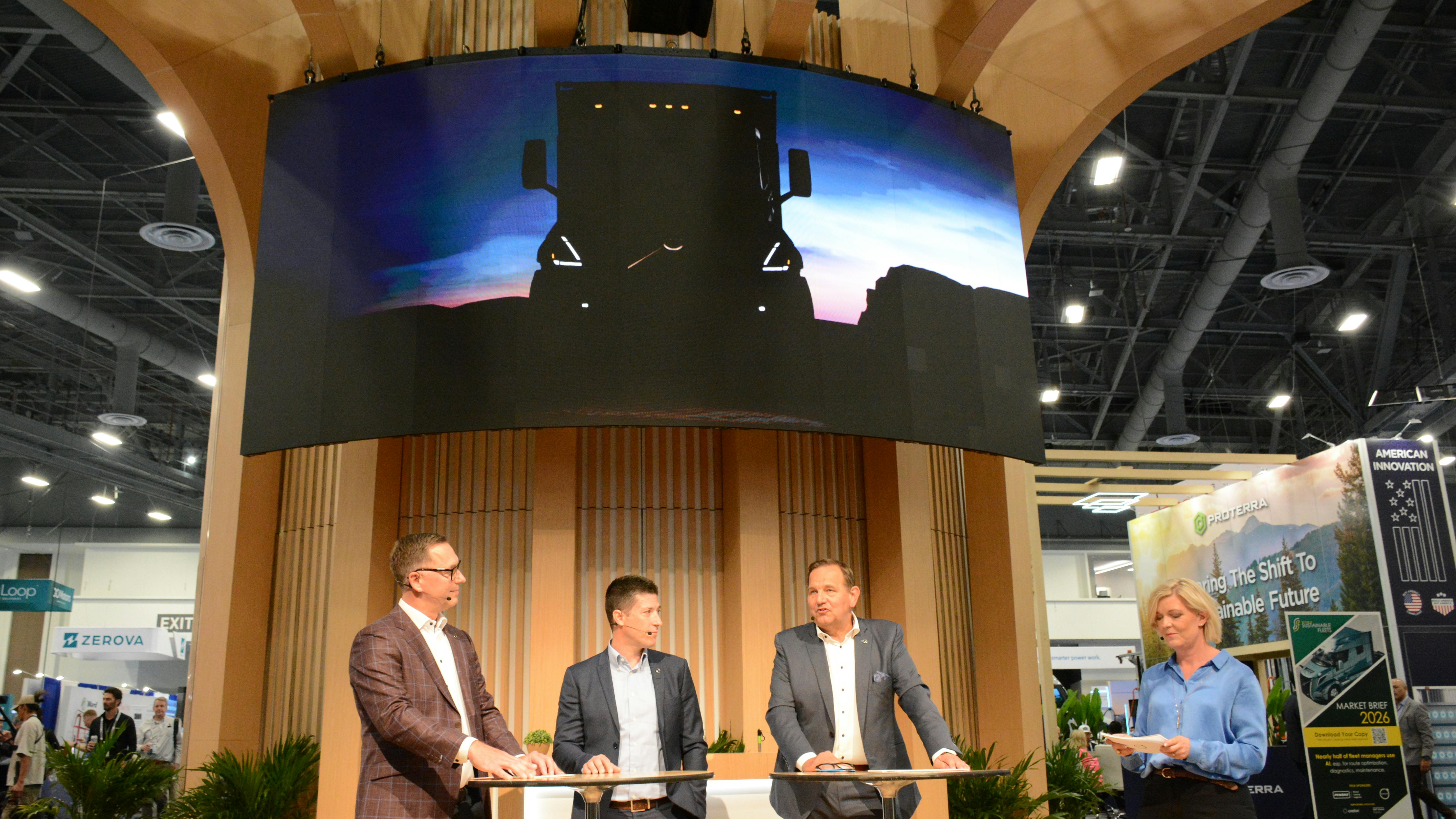 our Volvo executives standing on a stage at ACT Expo beneath a large digital screen displaying the yellow silhouette of a new heavy-duty vocational truck.