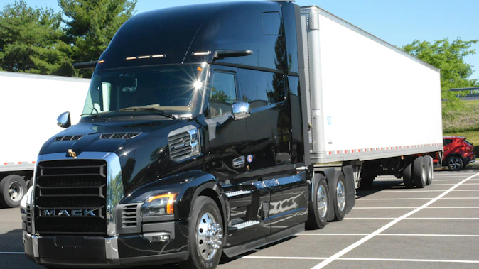 Three-quarter front view of a shiny black Mack Pioneer long-haul tractor hooked to a white 53-foot dry van trailer, parked on a paved lot.