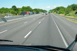 View from the driver's seat of a truck looking out through the windshield at a multi-lane highway (Interstate) with moderate traffic under a blue sky. The tips of the Mack Bulldog hood ornaments' ears are just barely visible, showing the wide view truck operates get inside the cab. View from the driver's seat of a truck looking out through the windshield at a multi-lane highway (Interstate) with moderate traffic under a blue sky. The tips of the Mack Bulldog hood ornaments' ears are just barely visible, showing the wide view truck operates get inside the cab.