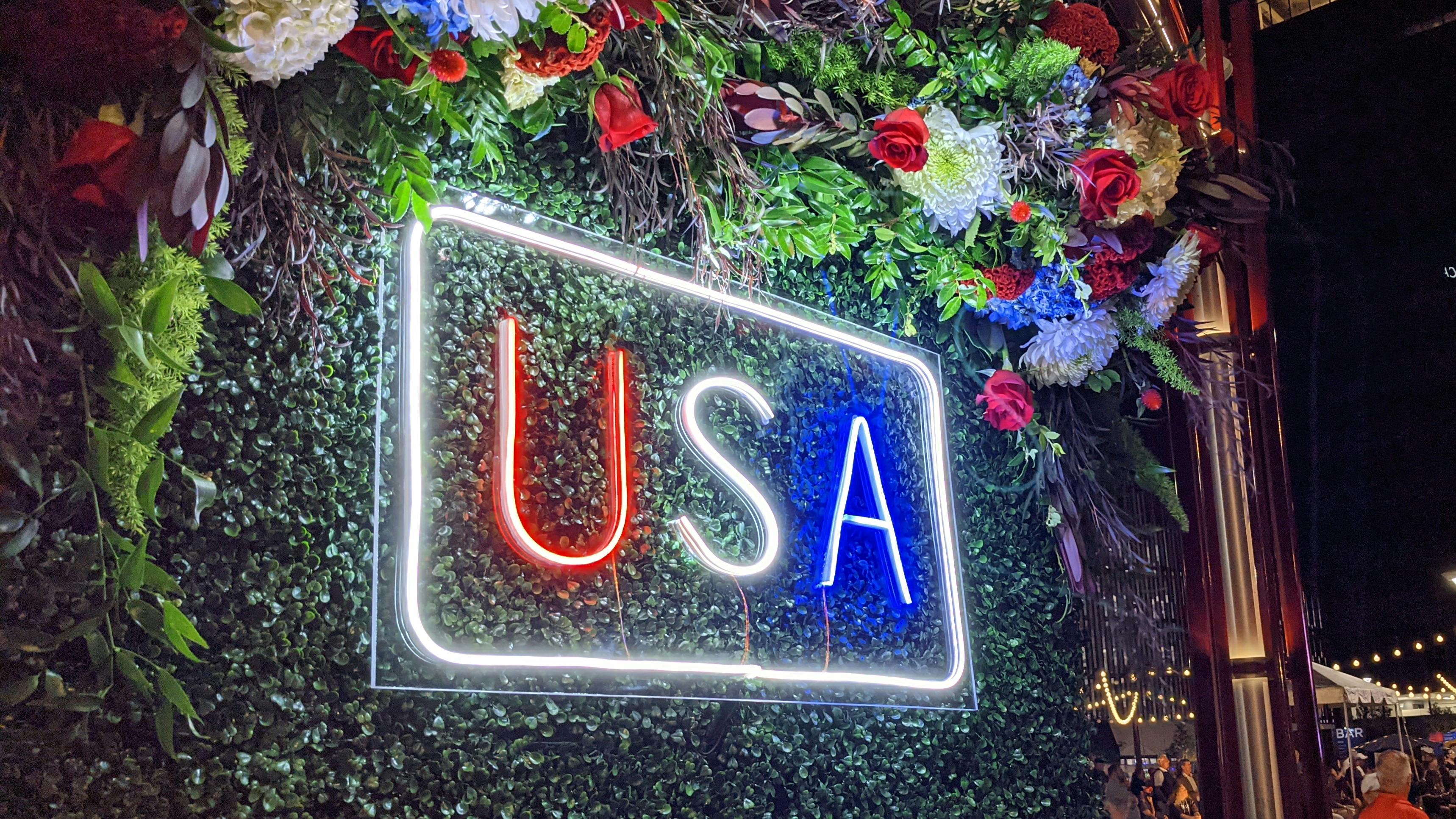 A glowing neon sign displaying 'USA' in red, white, and blue letters against a green foliage wall at night, surrounded by floral arrangements.