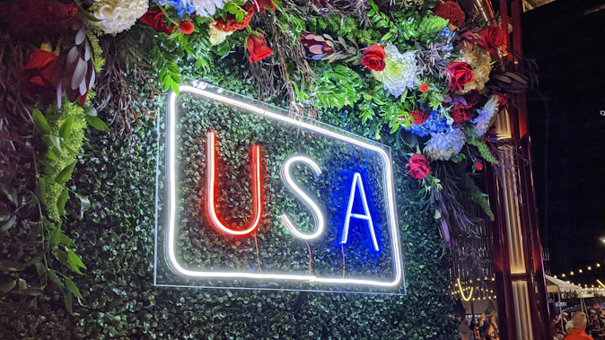 A glowing neon sign displaying 'USA' in red, white, and blue letters against a green foliage wall at night, surrounded by floral arrangements.
