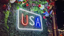 A glowing neon sign displaying 'USA' in red, white, and blue letters against a green foliage wall at night, surrounded by floral arrangements. A glowing neon sign displaying 'USA' in red, white, and blue letters against a green foliage wall at night, surrounded by floral arrangements.