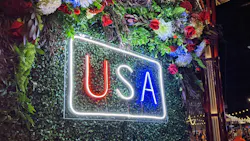 A glowing neon sign displaying 'USA' in red, white, and blue letters against a green foliage wall at night, surrounded by floral arrangements. A glowing neon sign displaying 'USA' in red, white, and blue letters against a green foliage wall at night, surrounded by floral arrangements.