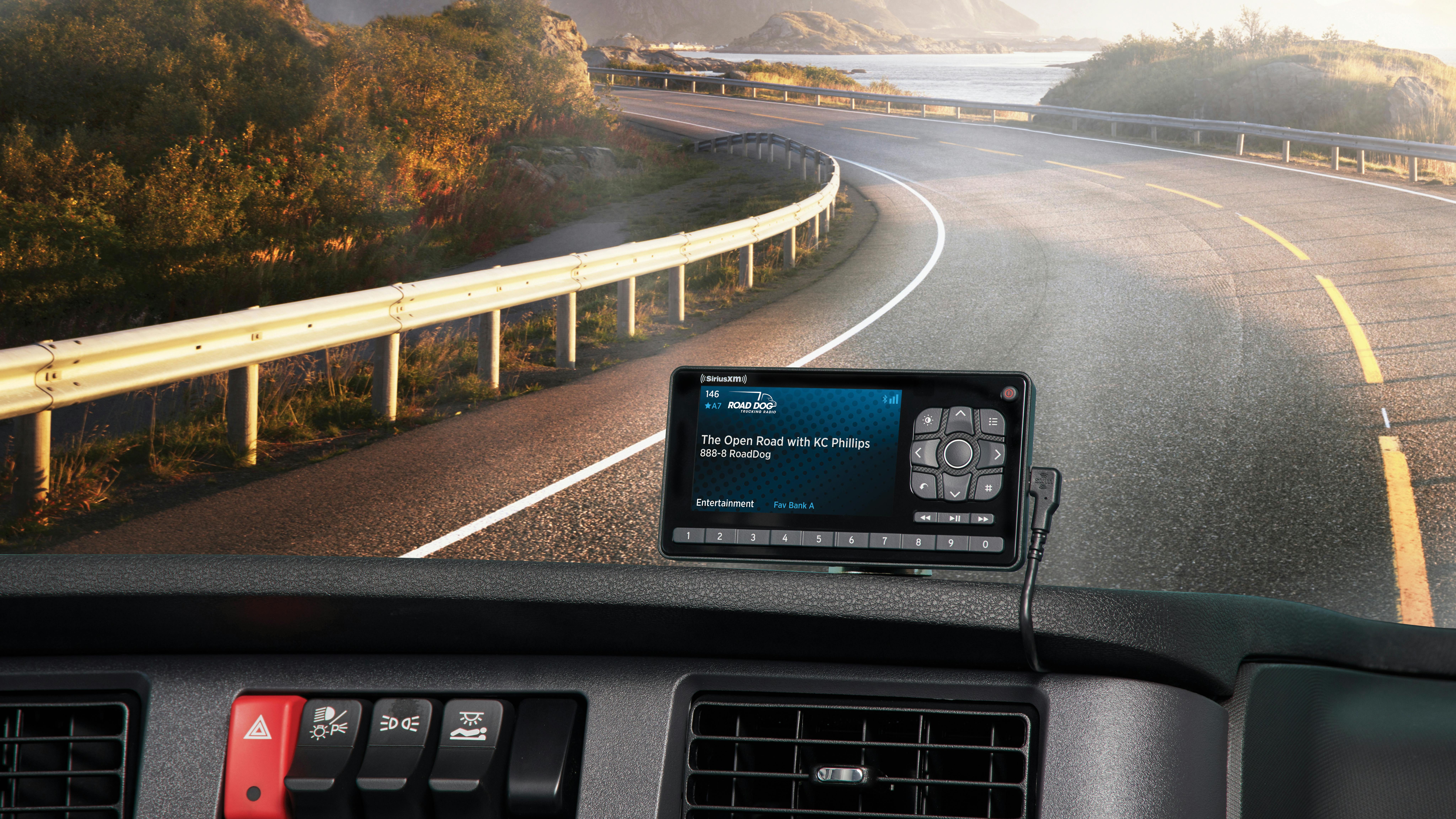 POV view from a commercial truck cab interior looking over the dashboard toward a winding scenic road by a lake and mountains; a satellite radio receiver displaying 'KC Phillips on Road Dog Trucking' is mounted on the dash.