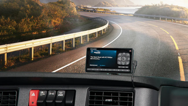 POV view from a commercial truck cab interior looking over the dashboard toward a winding scenic road by a lake and mountains; a satellite radio receiver displaying 'KC Phillips on Road Dog Trucking' is mounted on the dash.