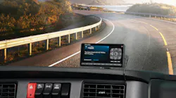 POV view from a commercial truck cab interior looking over the dashboard toward a winding scenic road by a lake and mountains; a satellite radio receiver displaying 'KC Phillips on Road Dog Trucking' is mounted on the dash.