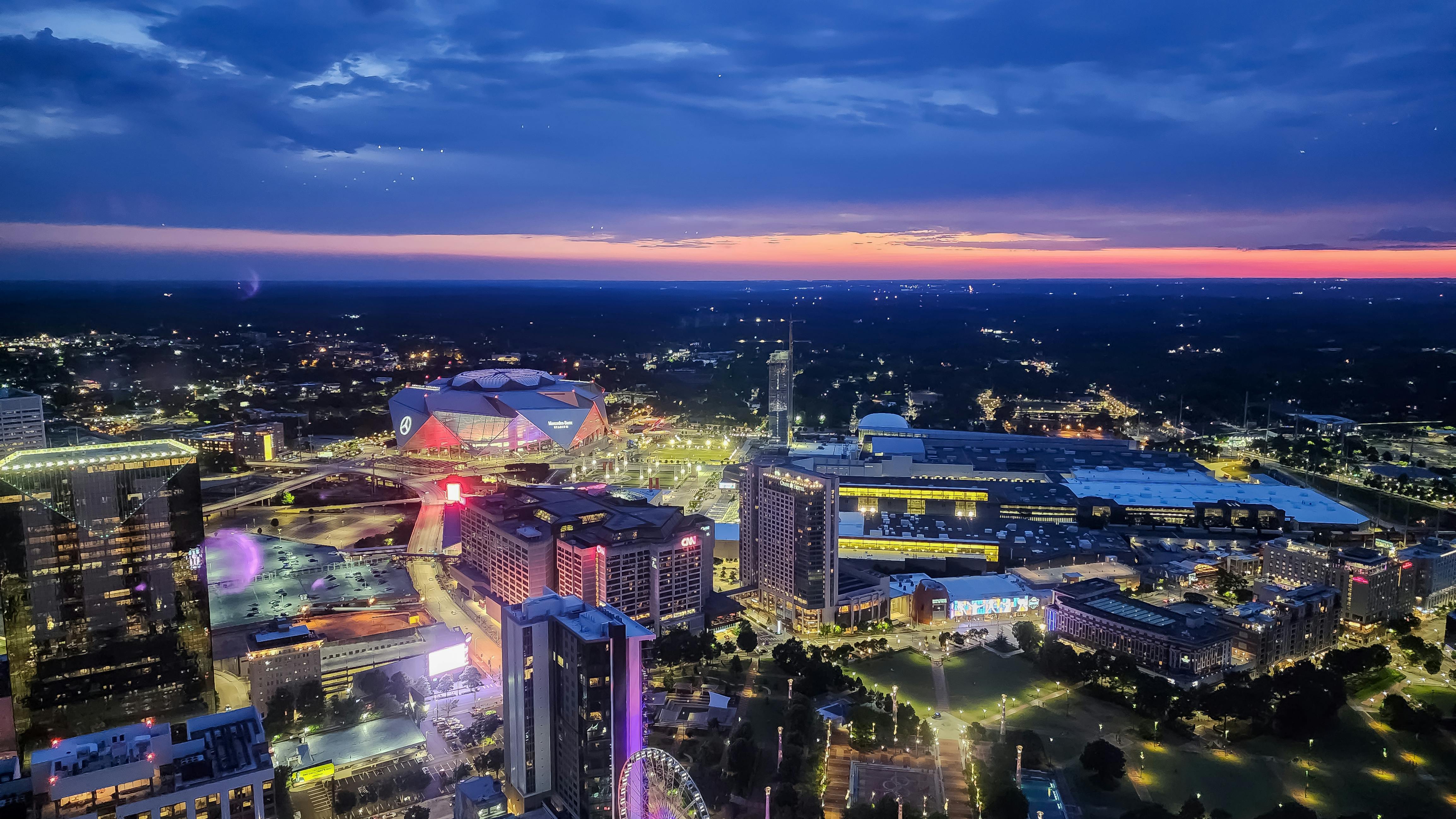 Aerial view of the Atlanta skyline at sunset with Mercedes-Benz Stadium, illuminated city streets, and surrounding buildings.