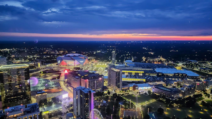 Aerial view of the Atlanta skyline at sunset with Mercedes-Benz Stadium, illuminated city streets, and surrounding buildings.