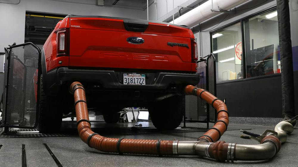 Rear view of a red Ford F-150 Raptor pickup truck strapped onto a dynamometer in a testing garage, with heavy-duty exhaust hoses connected to its dual tailpipes.