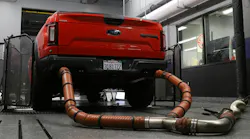 Rear view of a red Ford F-150 Raptor pickup truck strapped onto a dynamometer in a testing garage, with heavy-duty exhaust hoses connected to its dual tailpipes.