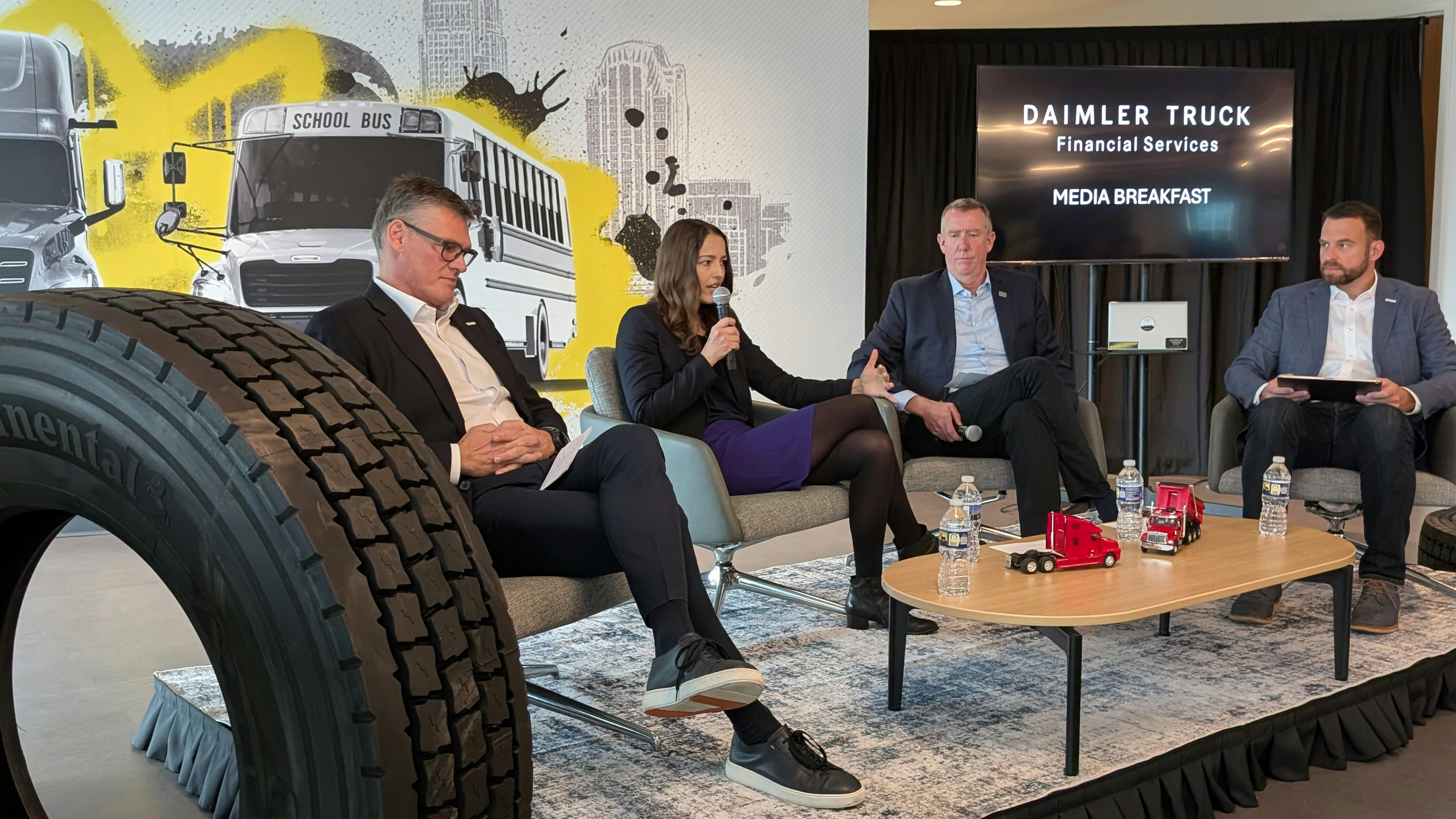 Four Daimler Truck executives sit on a stage during a media briefing in Charlotte in front of a backdrop showing Freightliner trucks and a school bus.