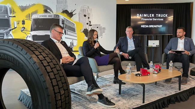 Four Daimler Truck executives sit on a stage during a media briefing in Charlotte in front of a backdrop showing Freightliner trucks and a school bus.